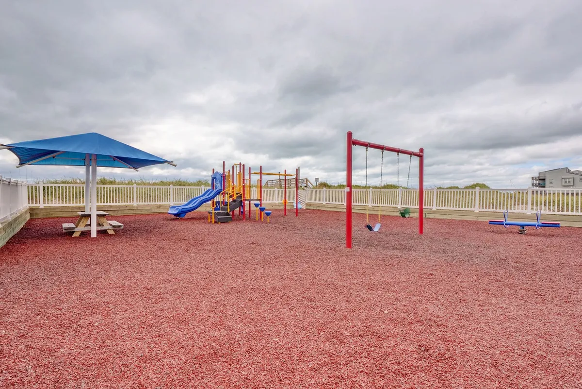 A lively outdoor playground with swings, play equipment, and a picnic area, bordered by a safety fence. The atmosphere is joyful and communal, inviting children and families to gather and play.