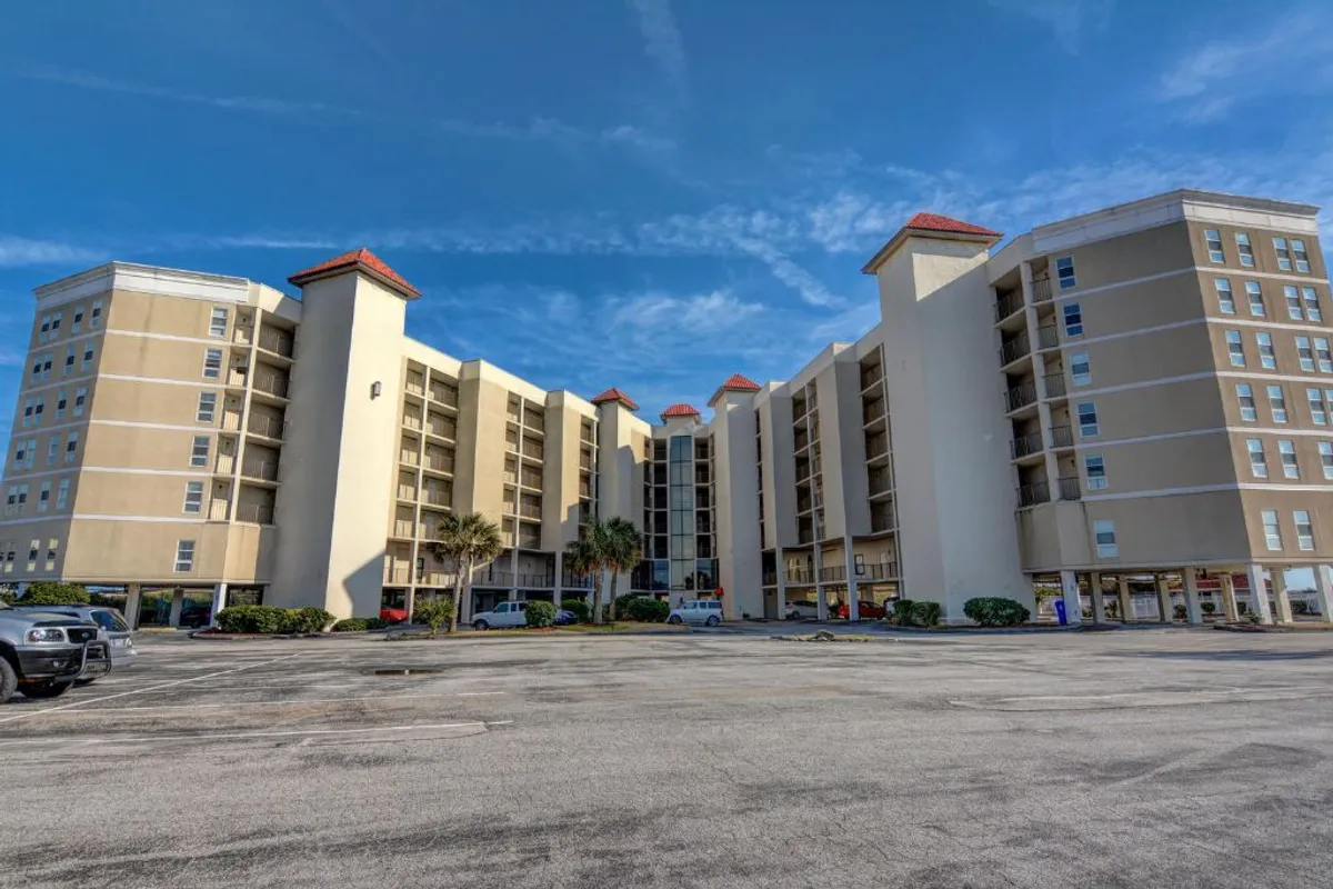 The resort's modern exterior featuring distinctive red-roof accents, palm trees, and a welcoming entrance area. The image captures sunny weather and a sense of arrival, suggesting the start of a fun, memorable group escape.