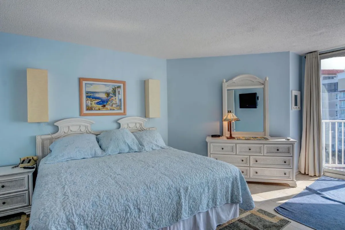 A serene master bedroom with a king-sized bed topped in a blue quilt, complemented by a decorative painting above the headboard and a dresser with a lamp. The room is filled with soft natural light, with clean lines and newly painted walls contributing to a fresh, tranquil atmosphere.
