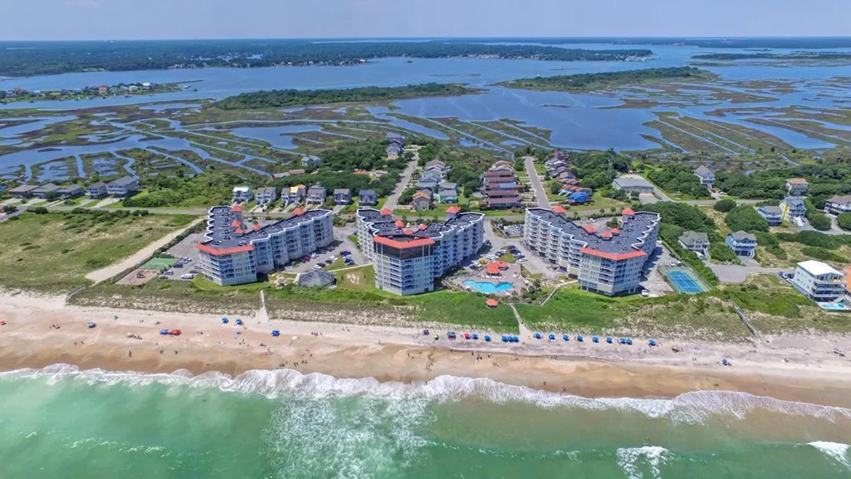 This image showcases beachfront buildings set against the backdrop of a beautiful sandy beach, a sparkling blue swimming pool, gentle ocean waves, and marshland beyond. The pool area is prominently in view, with easy access to the ocean just steps away.