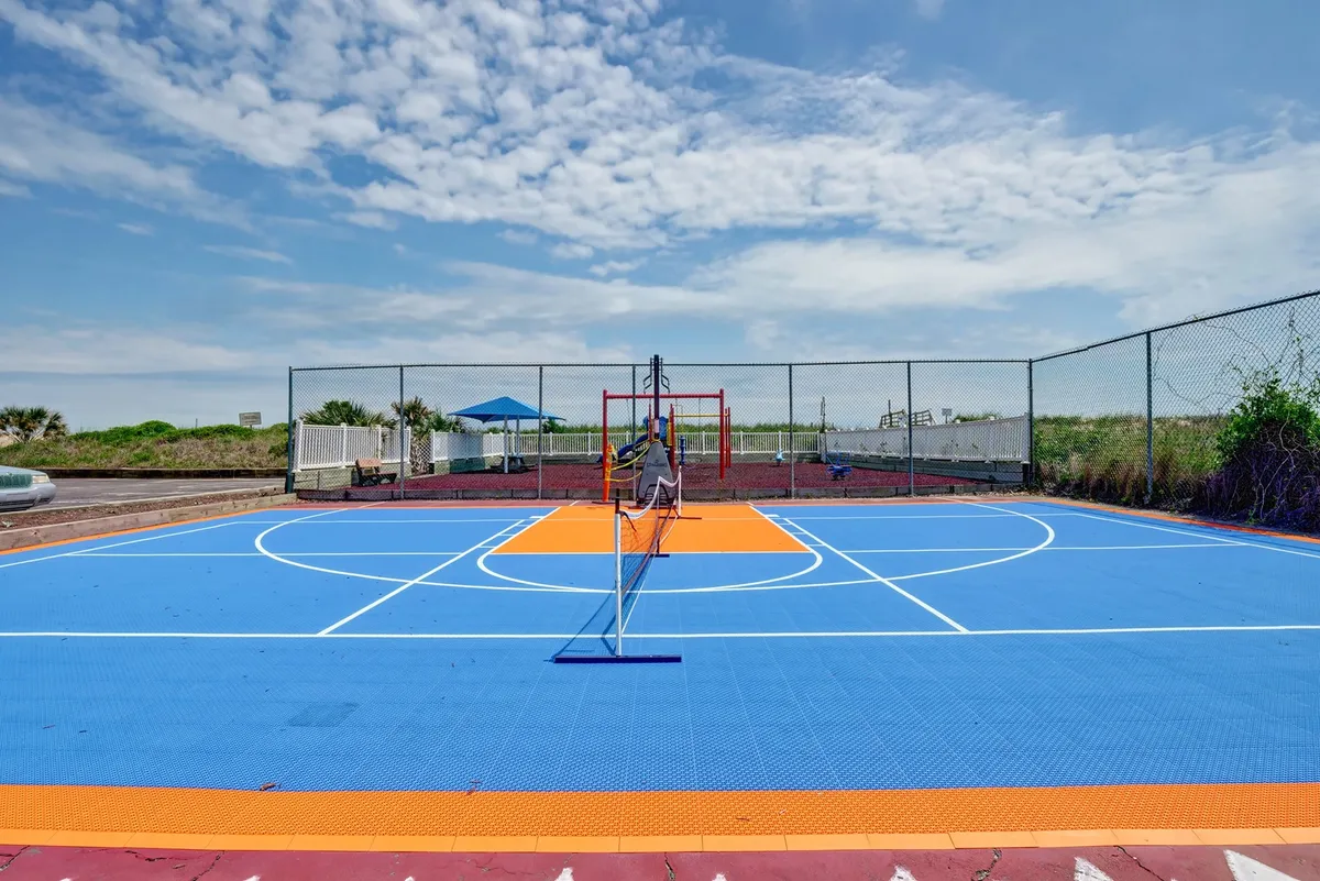 An energetic exterior scene showing a colorful sports court with a net, adjacent playground equipment, and green landscaping. People are engaged in basketball, tennis, and children are playing nearby, conveying a playful, community-focused vibe.