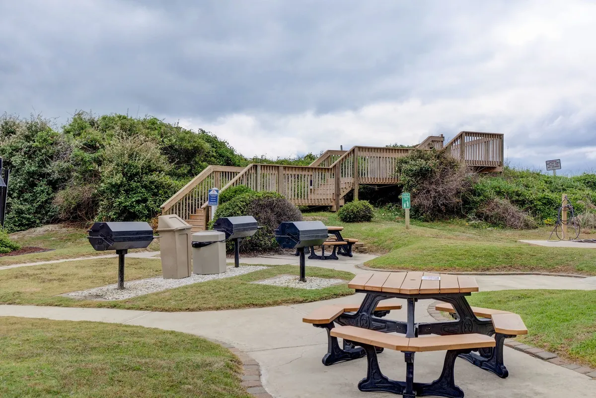 An exterior scene showing well-maintained picnic tables and grill stations surrounded by greenery and a stairway, creating a welcoming outdoor dining and socialization space. The area is clearly designed for guests to enjoy barbecues, picnics, and community gatherings, reinforcing the sense of shared amenities and outdoor activities.
