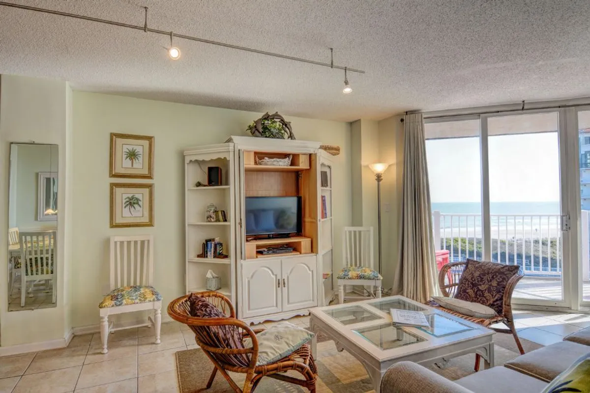 A serene bedroom setting with a cozy bed, large windows revealing panoramic ocean views, tasteful wall art, and a TV for relaxation. The ambiance is calm, welcoming, and intimate—ideal for couples seeking comfort and togetherness.