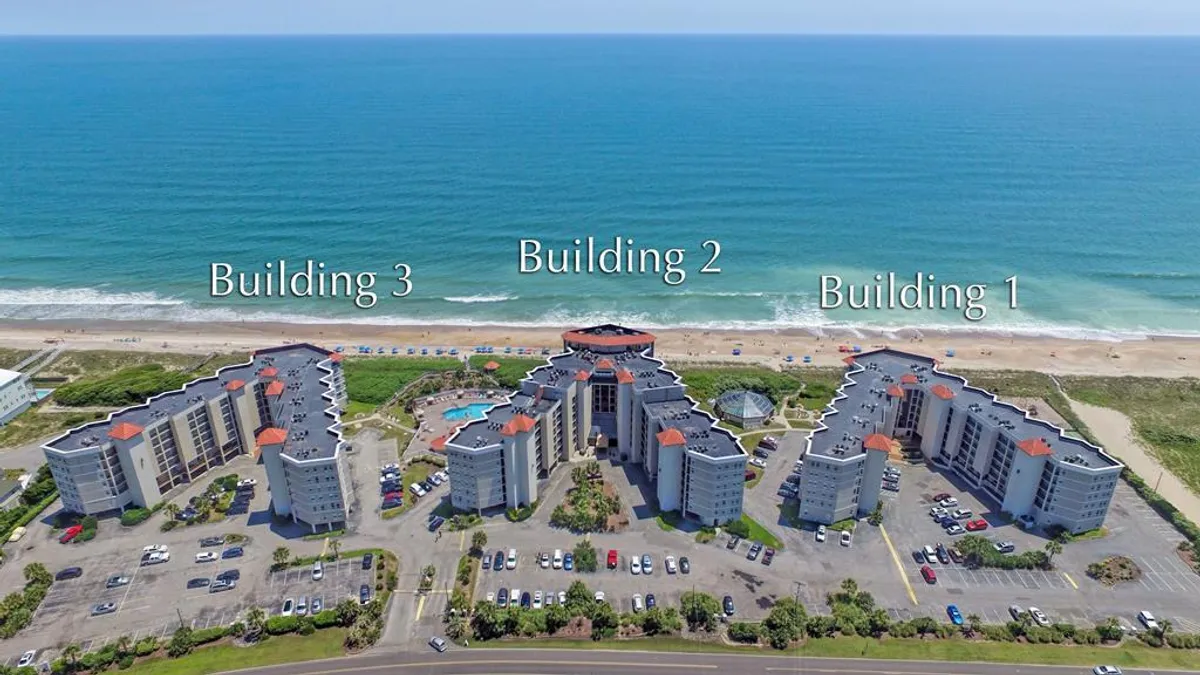 A wide exterior shot featuring the beachfront swimming pool area against the backdrop of three buildings. The pool is inviting, surrounded by lounge chairs and situated just steps from the sand, with balconies overlooking both the pool and the coastline. The scene conveys the ease of accessing both the beach and the pool, while also hinting at communal spaces for gatherings and relaxation.