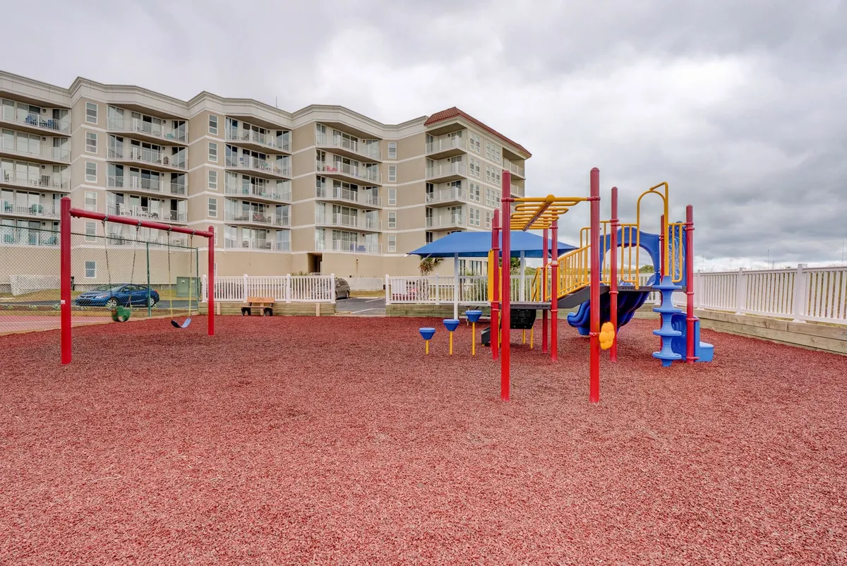 A joyful, colorful playground scene with swings and other play equipment set on red rubber mulch, positioned in front of an apartment-style building. The area is lively and clearly designed for children and family interaction, conveying a sense of fun, safety, and community.