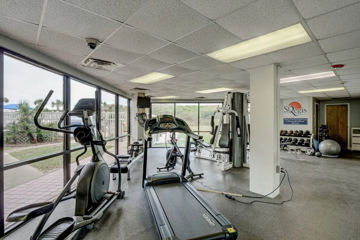 A well-lit fitness center featuring treadmills, elliptical machines, weight training equipment, and large windows letting in natural light. The scene includes people working out and engaging in group fitness activities.