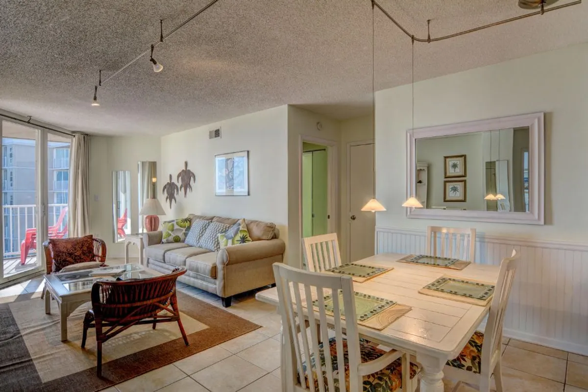 This image showcases a bright, open living area featuring a plush sofa with colorful cushions and a dining table set with placemats. Large windows flood the space with natural light, creating an inviting, airy atmosphere. The setup clearly highlights the seamless flow between the living and dining areas, perfect for gatherings and shared meals.