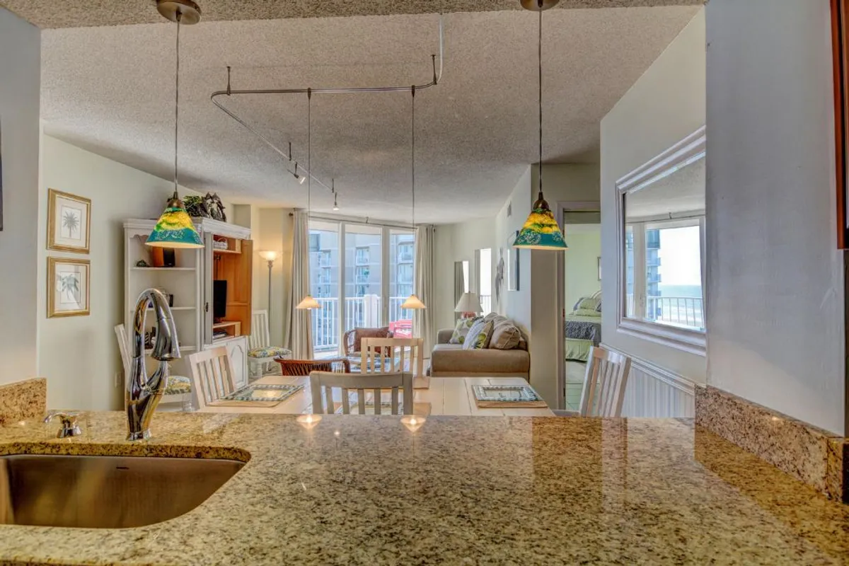 A welcoming interior scene featuring a granite countertop and a living area with a plush sofa. Hanging pendant lights provide warmth while large windows reveal a view beyond, hinting at a connection to the balcony. The arrangement suggests a space designed for dining, relaxing, and socializing.
