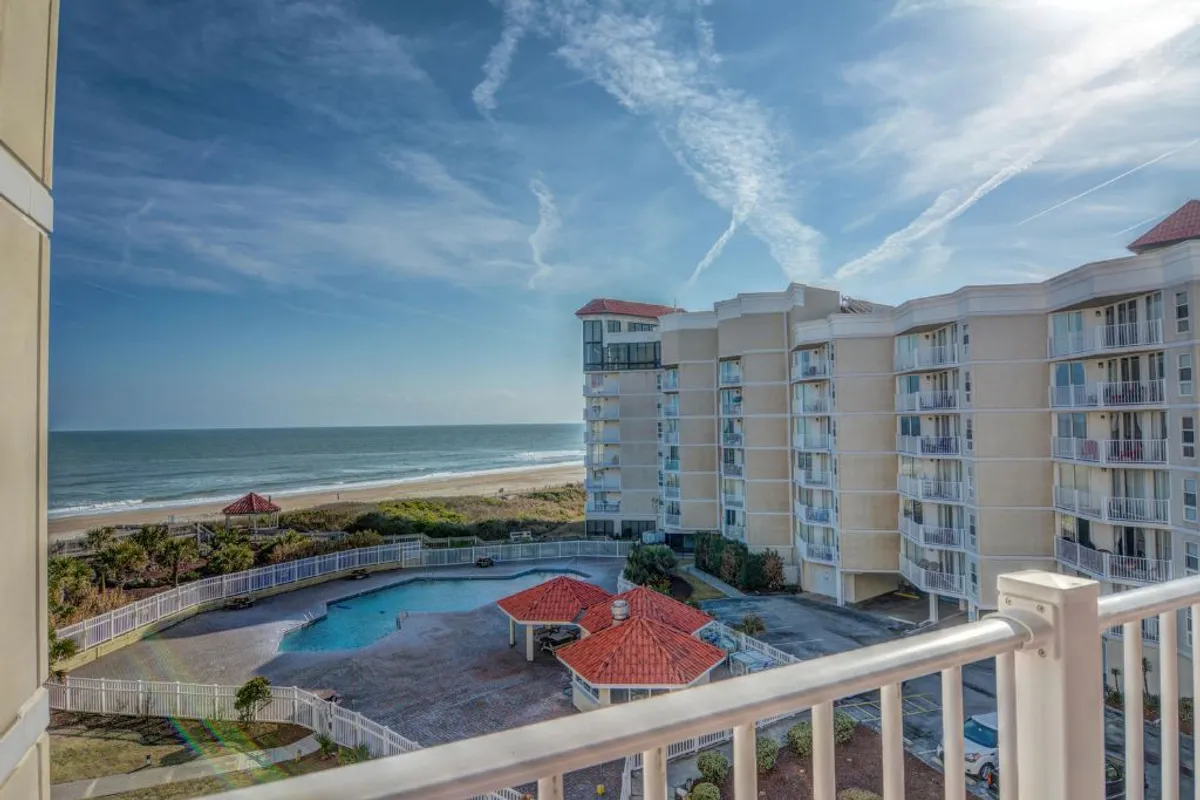 A sweeping view from a balcony shows the sparkling swimming pool below, red-roofed cabana, and wide ocean horizon. The scene includes the pool area, beach, and ocean—all visible at once, emphasizing the proximity of amenities.
